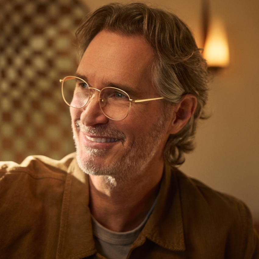 Man wearing gold-rimmed eyeglasses, sporting a brown jacket over a gray shirt, smiling.