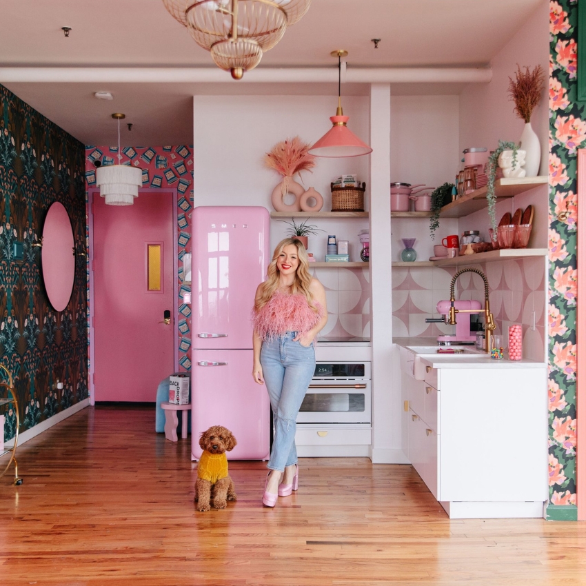 Woman in a pink feather top and jeans standing near a pink Smeg refrigerator with a brown dog in yellow sweater.