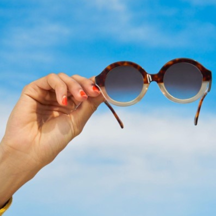 Hand holding round, tortoiseshell sunglasses with gradient lenses against a blue sky backdrop.