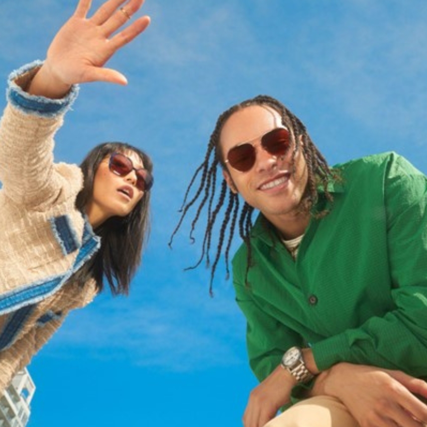 Two people wearing sunglasses, smiling, and posing against a bright blue sky background.