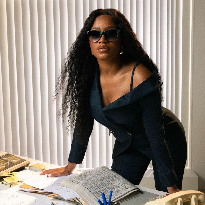 Woman in a professional blazer and sunglasses leaning over a desk cluttered with papers and office supplies.