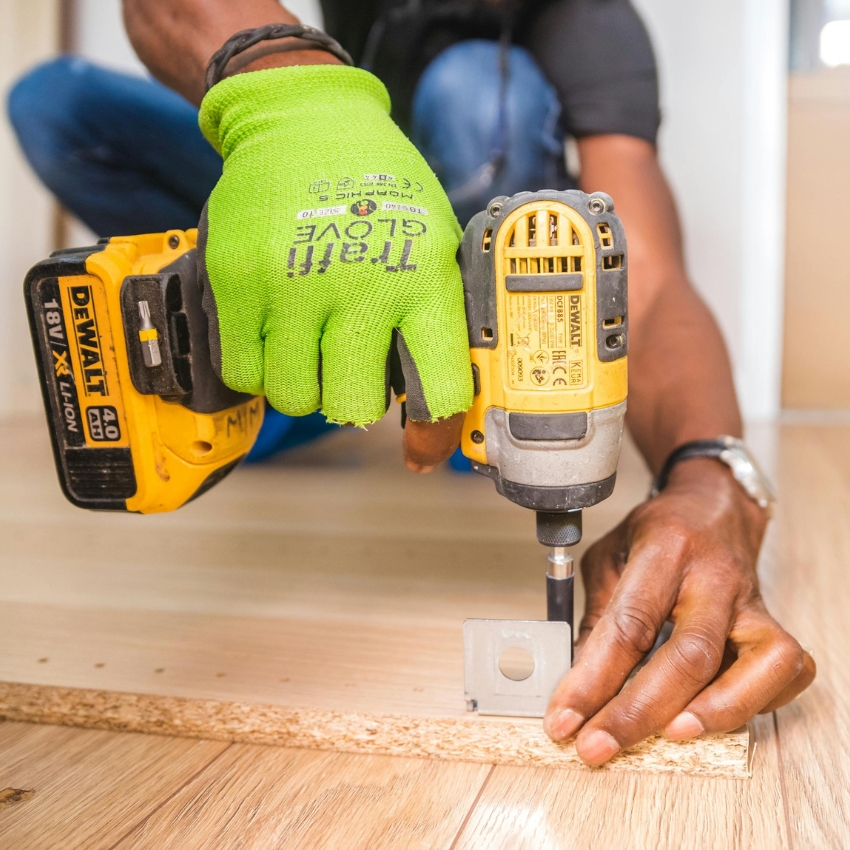 Dewalt cordless drill in use, held by a person wearing a green glove labeled "TRITON GLOVE ANSI 105-2016."