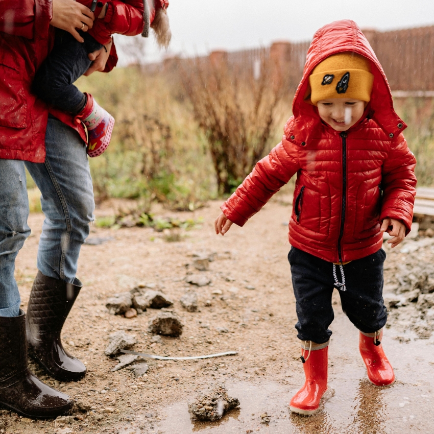 Child wearing red rain boots, a red puffer jacket, and a yellow beanie walking in mud. Adult in black boots holding another child's leg.