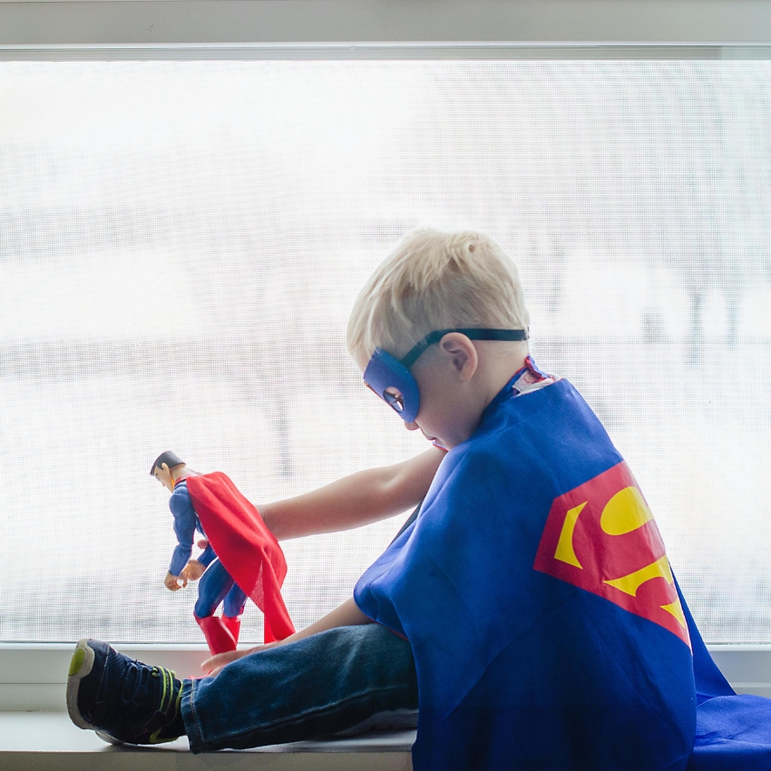 Child wearing a blue superhero cape and mask, playing with a Superman action figure.