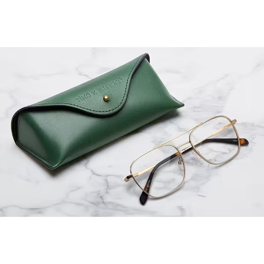 Green eyeglass case with "TIRO x PARISSP" text, beside gold-framed glasses on marble surface.