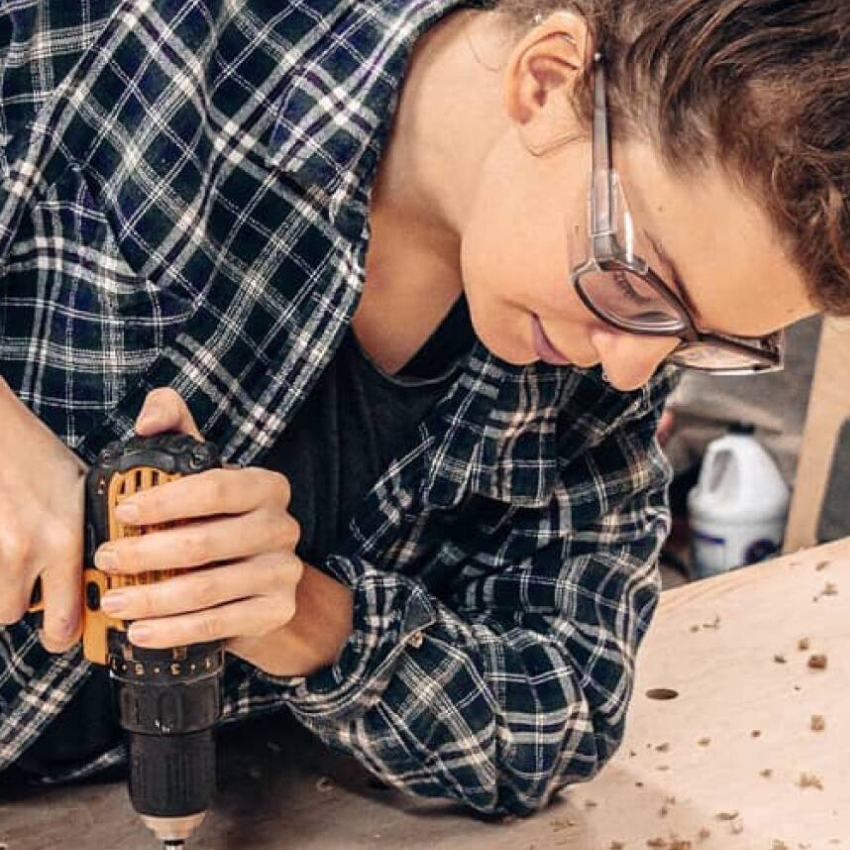 Person using a cordless power drill on a wooden surface while wearing safety glasses and a plaid shirt.