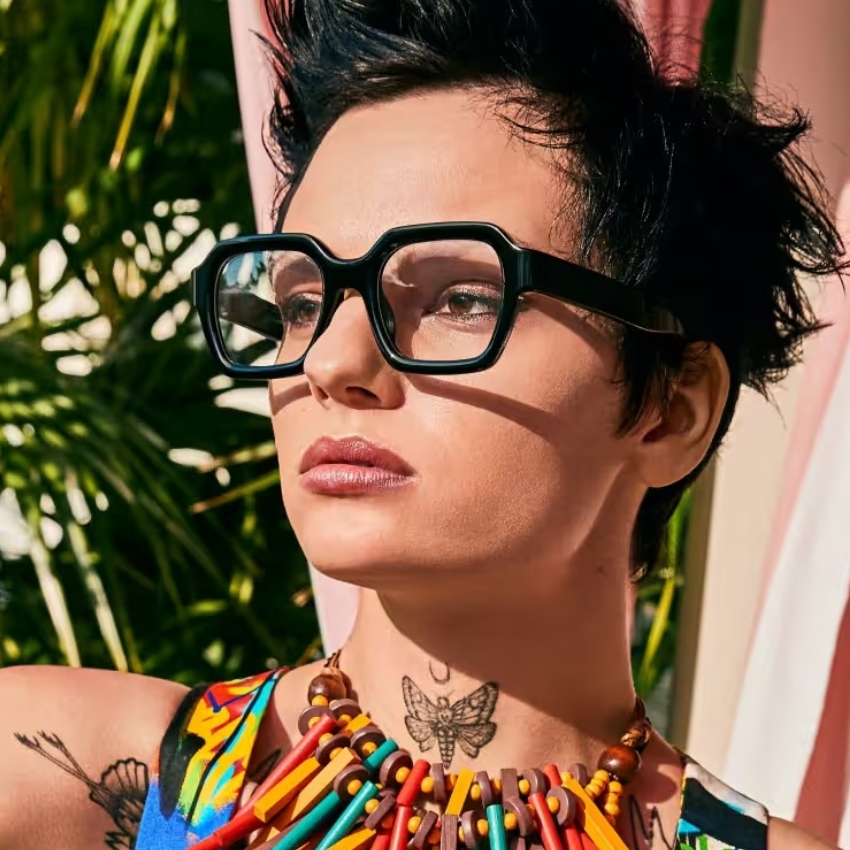 Person wearing oversized black glasses and a colorful necklace of various wooden pencils arranged in a pattern.