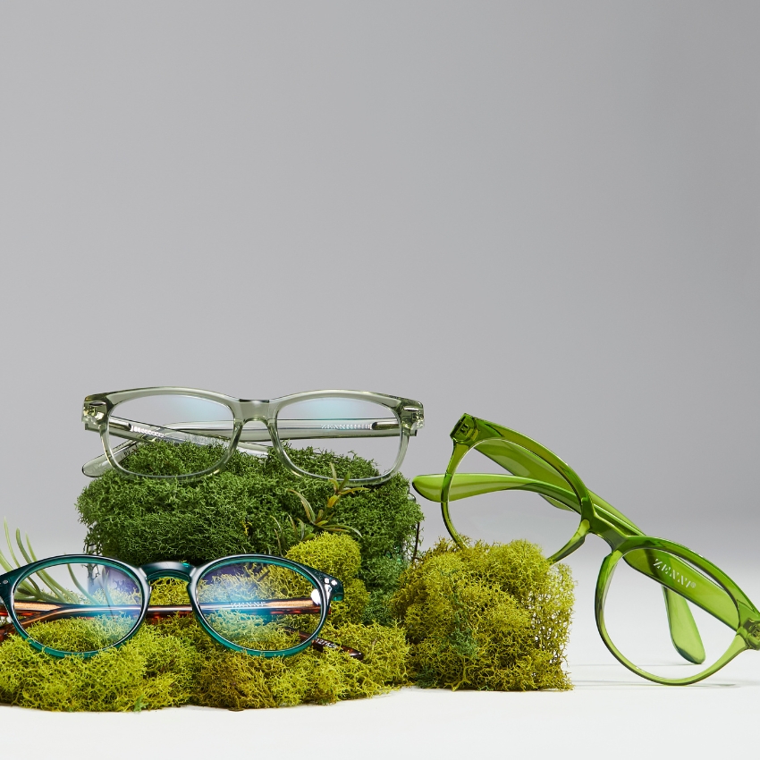 Three pairs of green eyeglasses displayed on mossy cushions. One pair has the name Zeal Optics on the side.