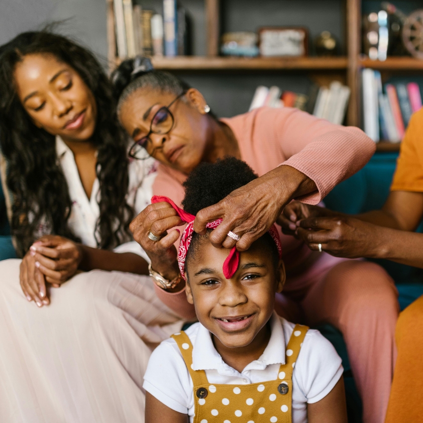 Elderly woman tying a red bandana on a smiling girl's hair, who is wearing a yellow polka dot dress.