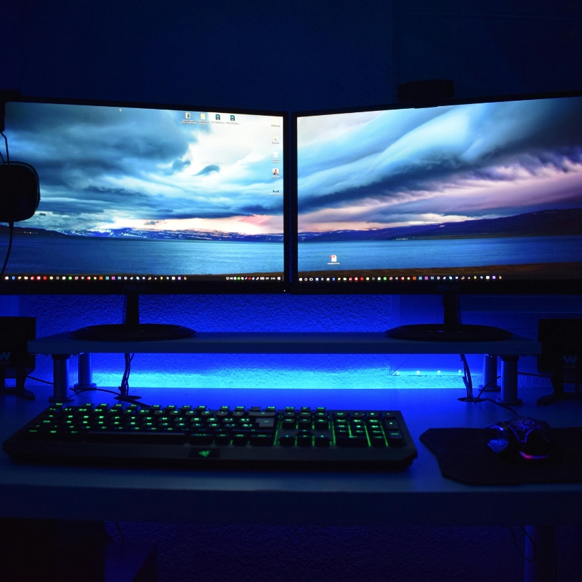 Dual monitors displaying a scenic landscape, backlit keyboard with green lights, and a gaming mouse.