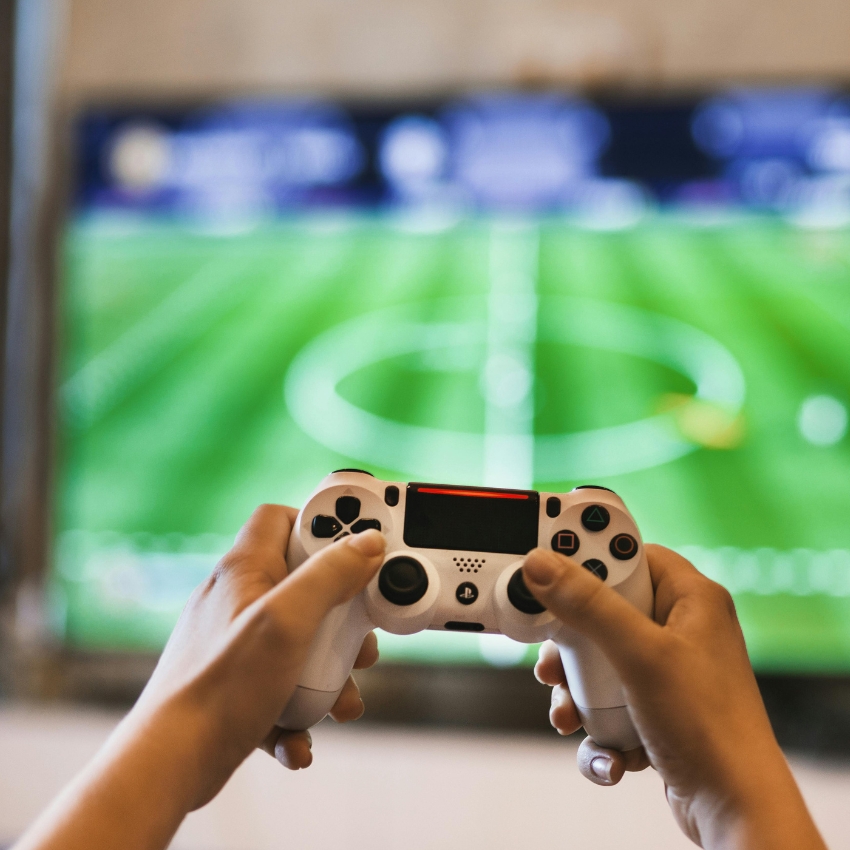 Hands holding a white gaming controller in front of a TV displaying a soccer video game.