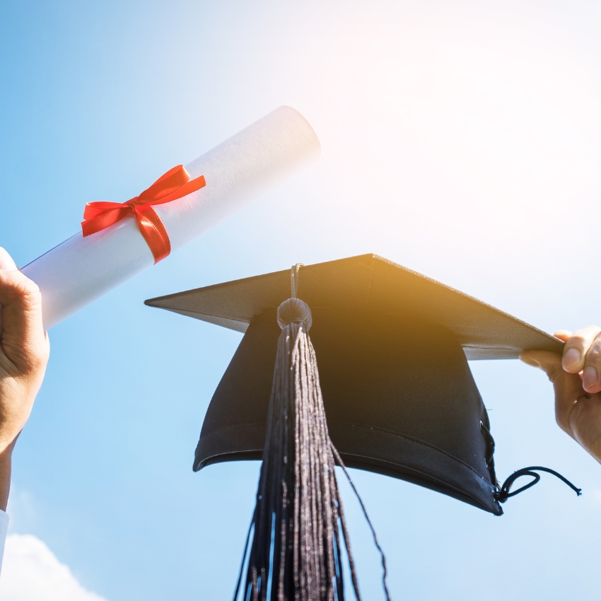 Graduation cap with tassel and diploma tied with a red ribbon held against a bright sky.