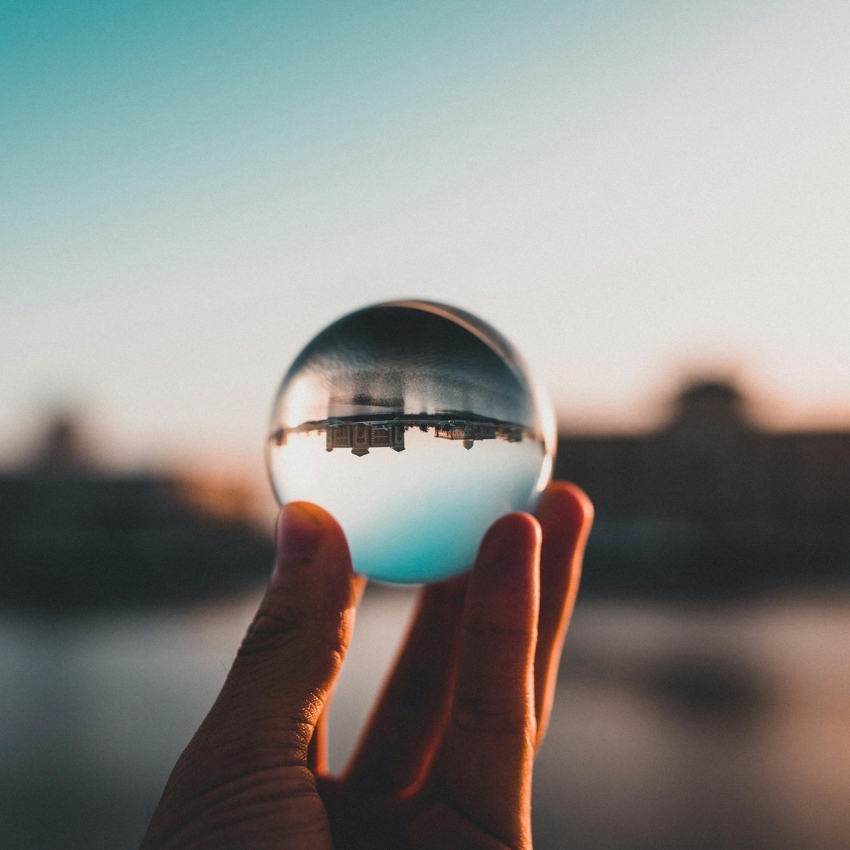 Hand holding a clear glass sphere with an inverted landscape inside.