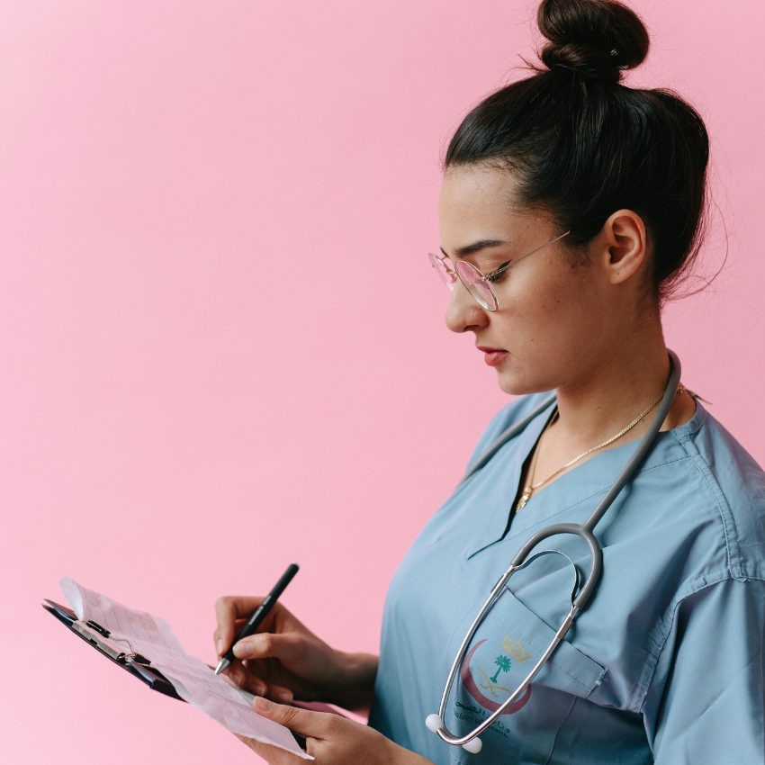 Nurse in blue scrubs with stethoscope, writing on a clipboard against a pink background.