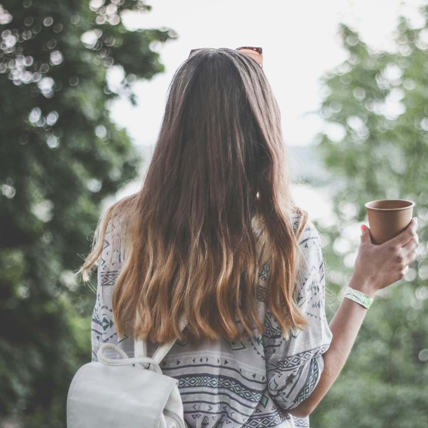 Woman with ombre hair, carrying a white backpack, holds a brown disposable cup while wearing a patterned top.