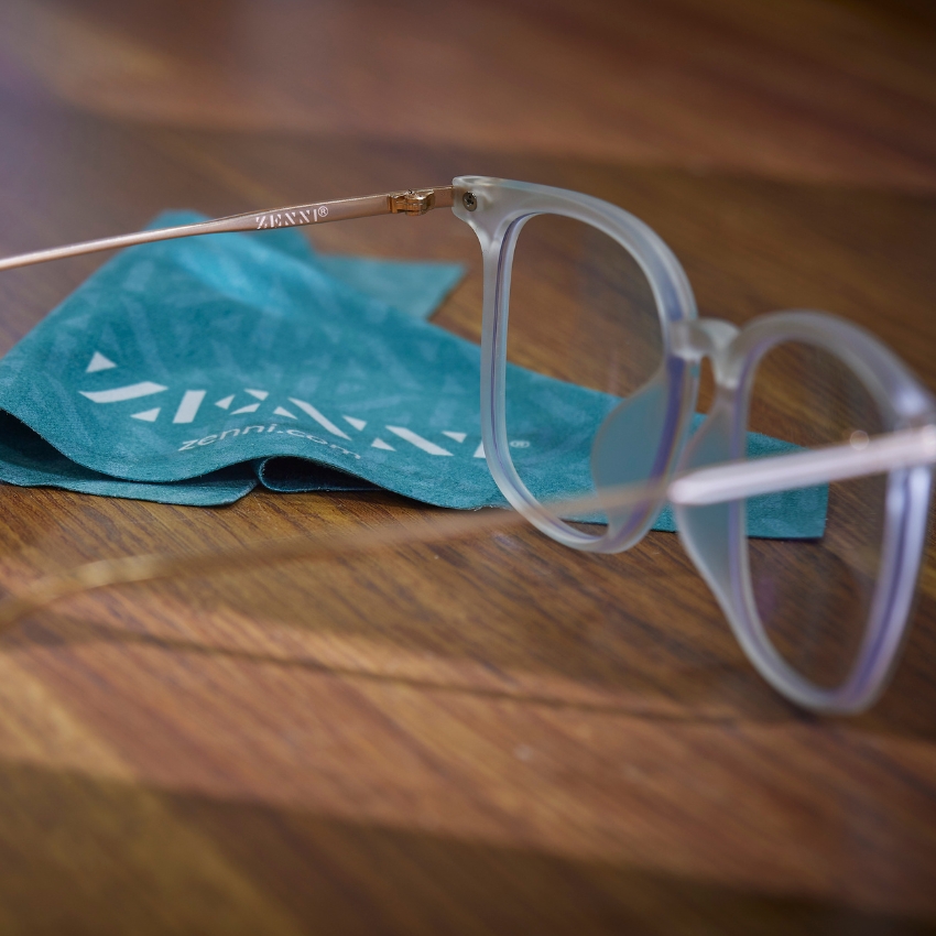 Clear-framed glasses with "ZENNI" on the temples, resting on a blue cleaning cloth that reads "zenni.com."