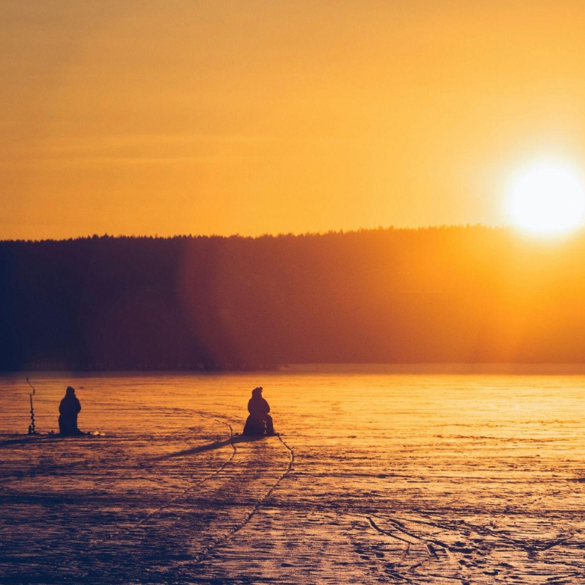 Two people ice fishing at sunrise on a frozen lake.