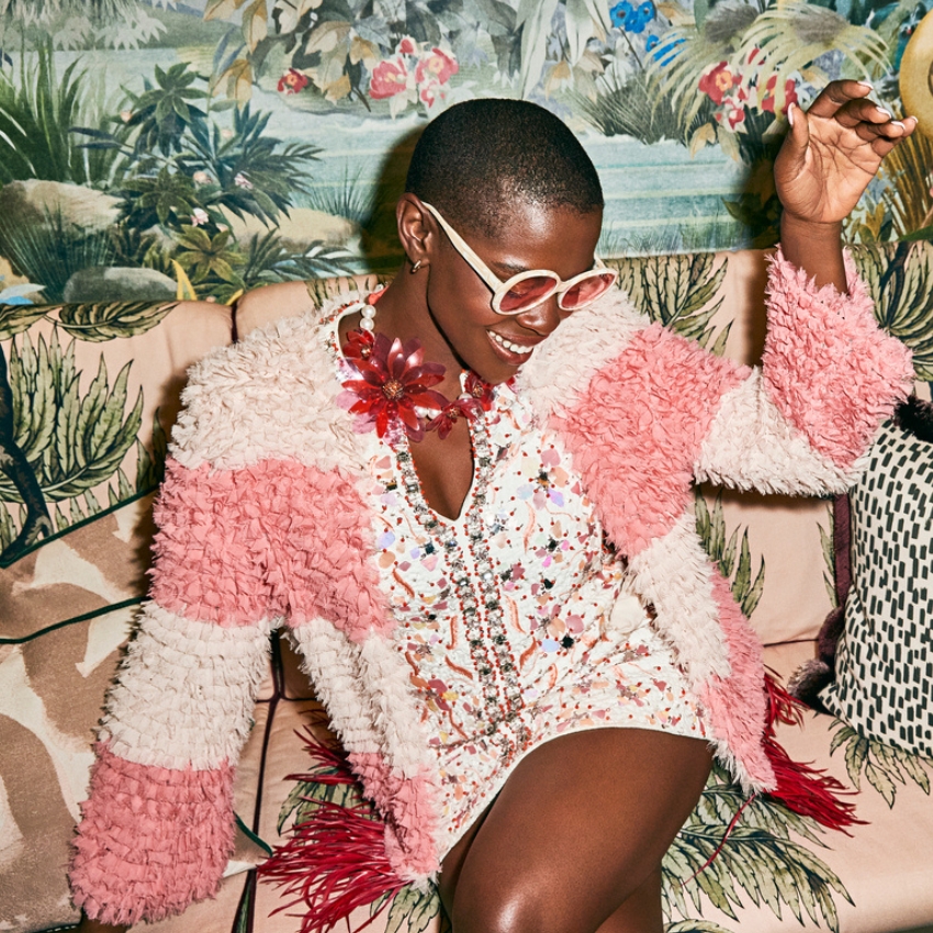 Woman wearing pink and white fluffy jacket, floral necklace, and sequin dress with red fringe.