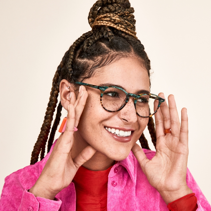 Woman wearing green tortoiseshell glasses, pink shirt, and red undershirt, smiling with hands raised.