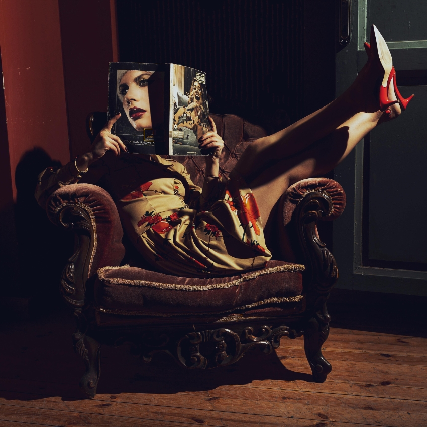 Woman in a floral dress reading a fashion magazine, wearing white heels with red soles, sitting on a vintage chair.