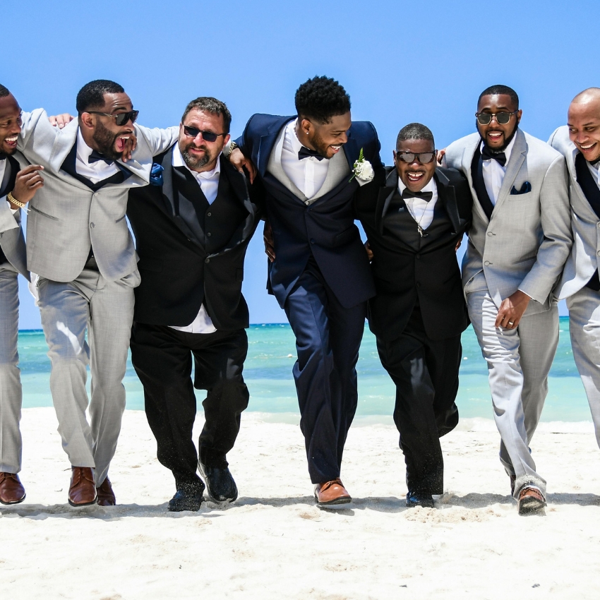 Groomsmen in suits and tuxedos smiling and walking on the beach with arms around each other.