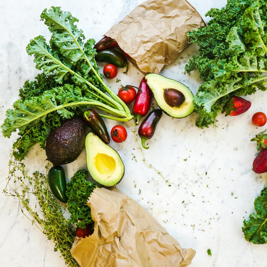 Avocados, kale, cherry tomatoes, and various peppers on a marble surface with torn brown paper bags.