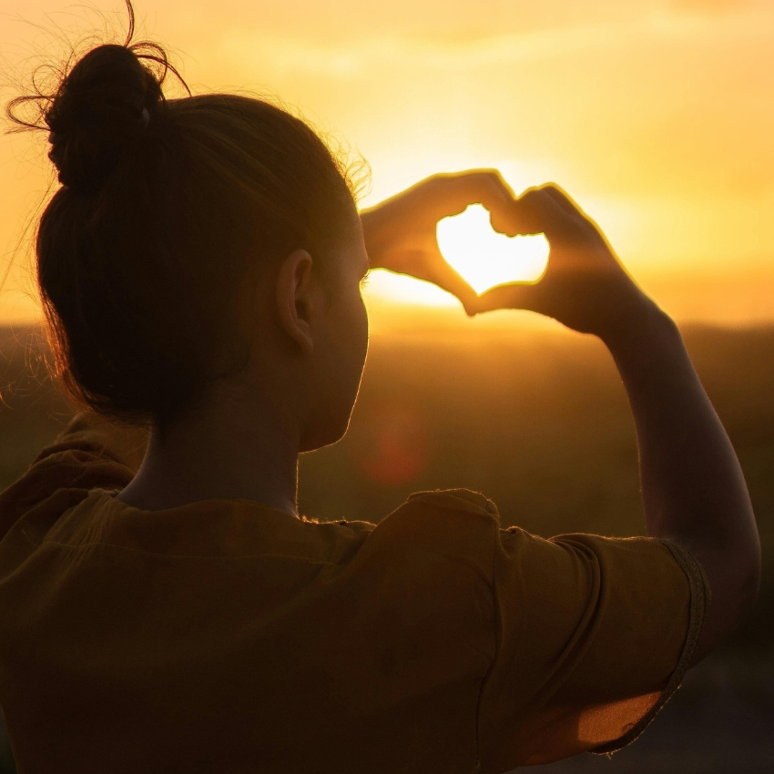 Person making a heart shape with their hands against a sunset.