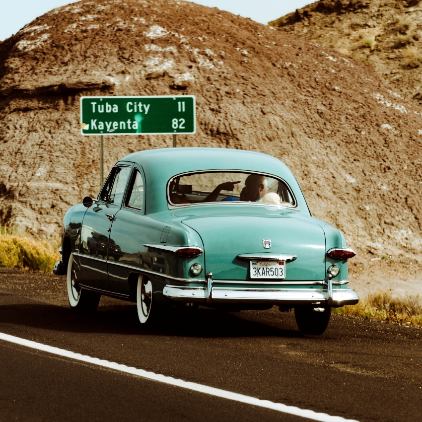 Turquoise vintage car driving on a road. Road sign reads "Tuba City 11 Kayenta 82." License plate 3KAR503.