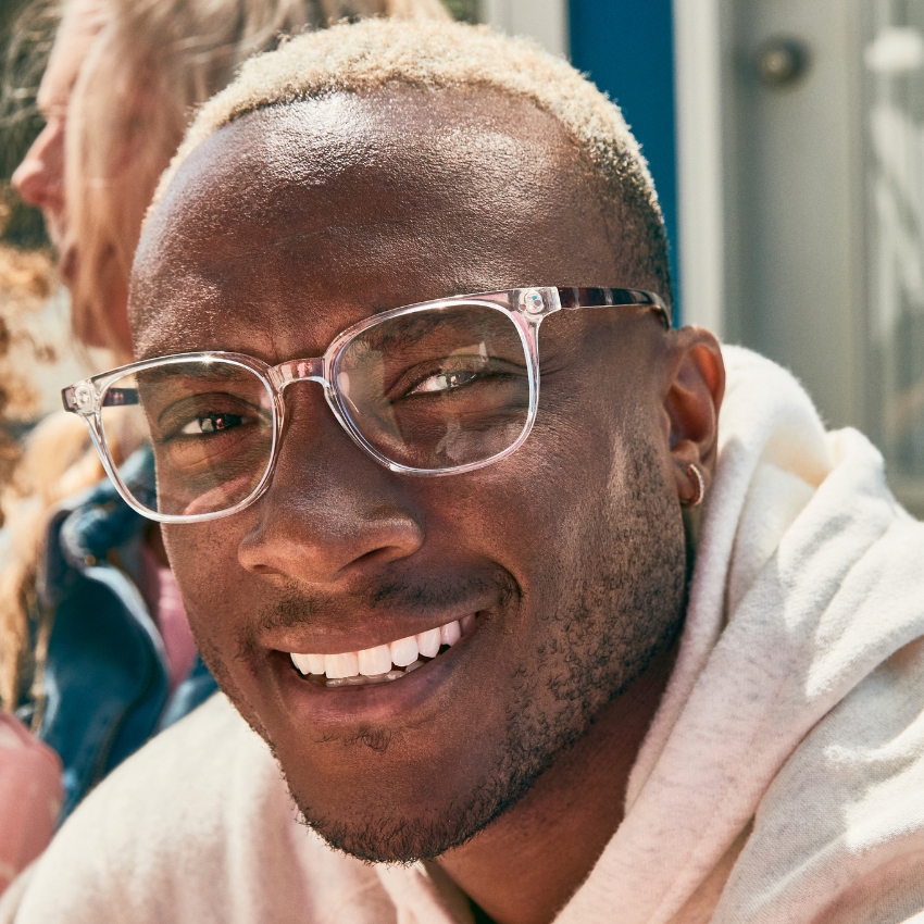 Man wearing clear-framed glasses and a light-colored hoodie, smiling outdoors.