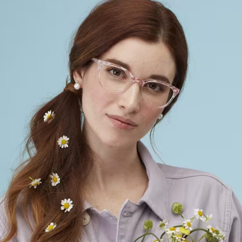 Woman wearing stylish clear pink eyeglasses, holding a bouquet of flowers, with daisies in her hair.