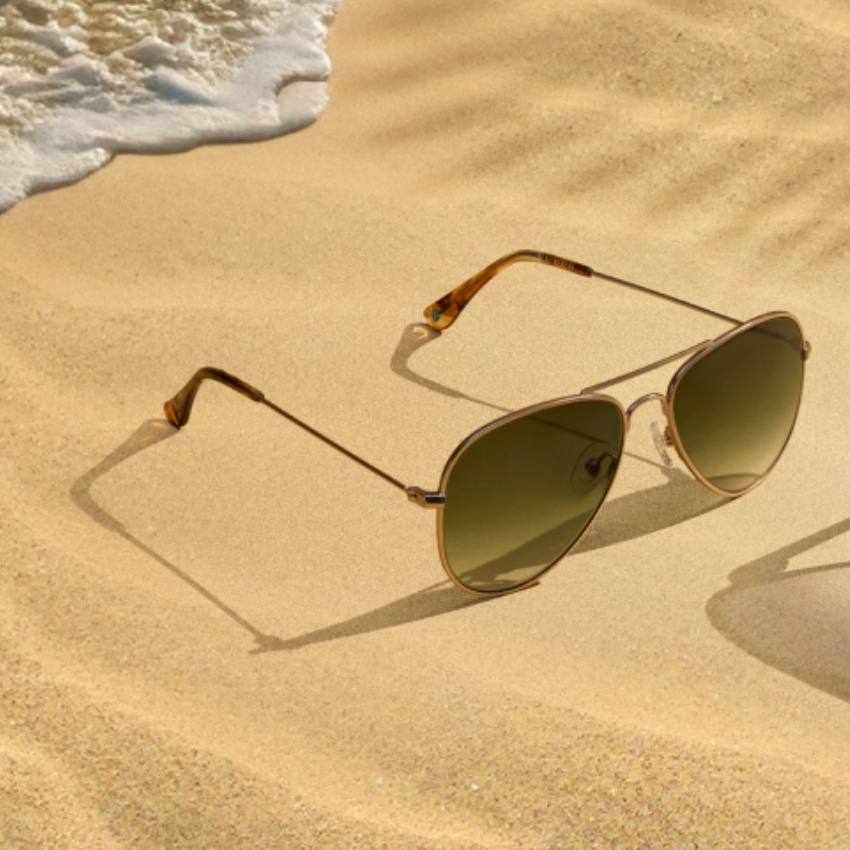 Gold-framed aviator sunglasses with dark lenses on sandy beach.