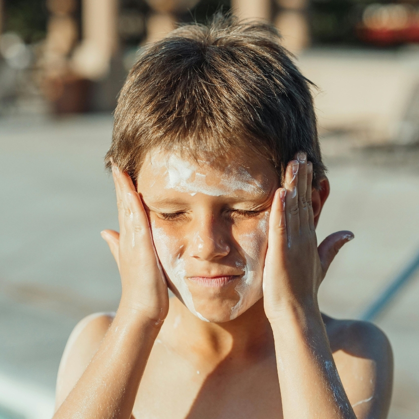 Child applying sunscreen to face, covering cheeks and forehead with eyes closed.