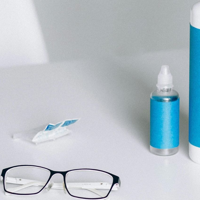 Eyeglasses with a blue frame; beside a blue-labeled eye drop bottle and a lens cleaning cloth.