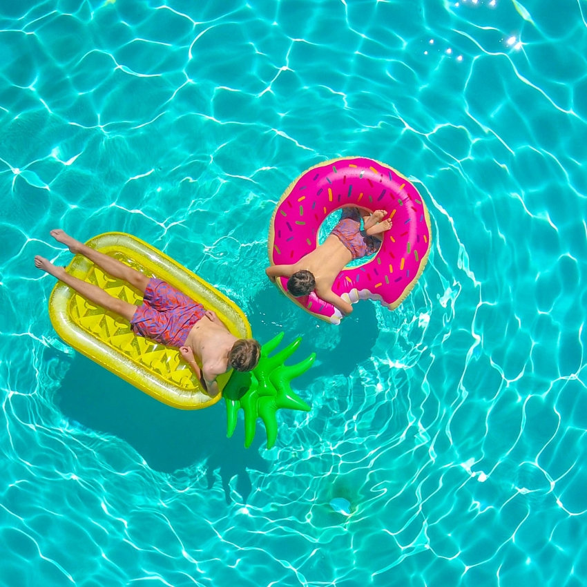 Two children in swimsuits float on pool inflatables: a pineapple lounge and a pink donut ring.