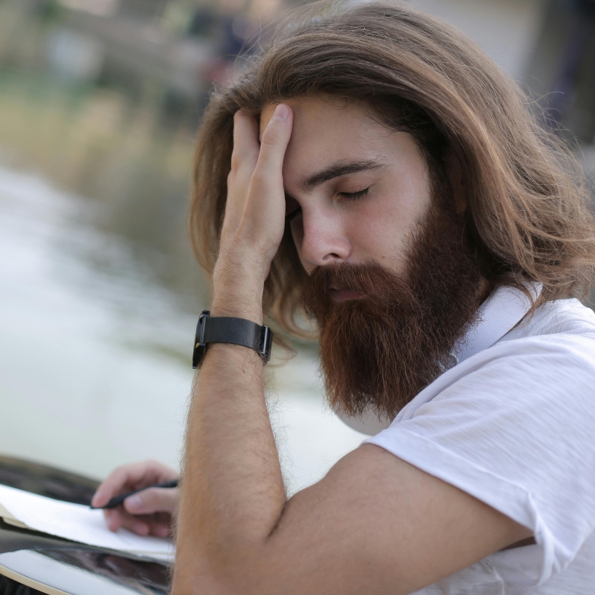 Person with long hair and beard, wearing a white shirt and wristwatch, resting hand on forehead.