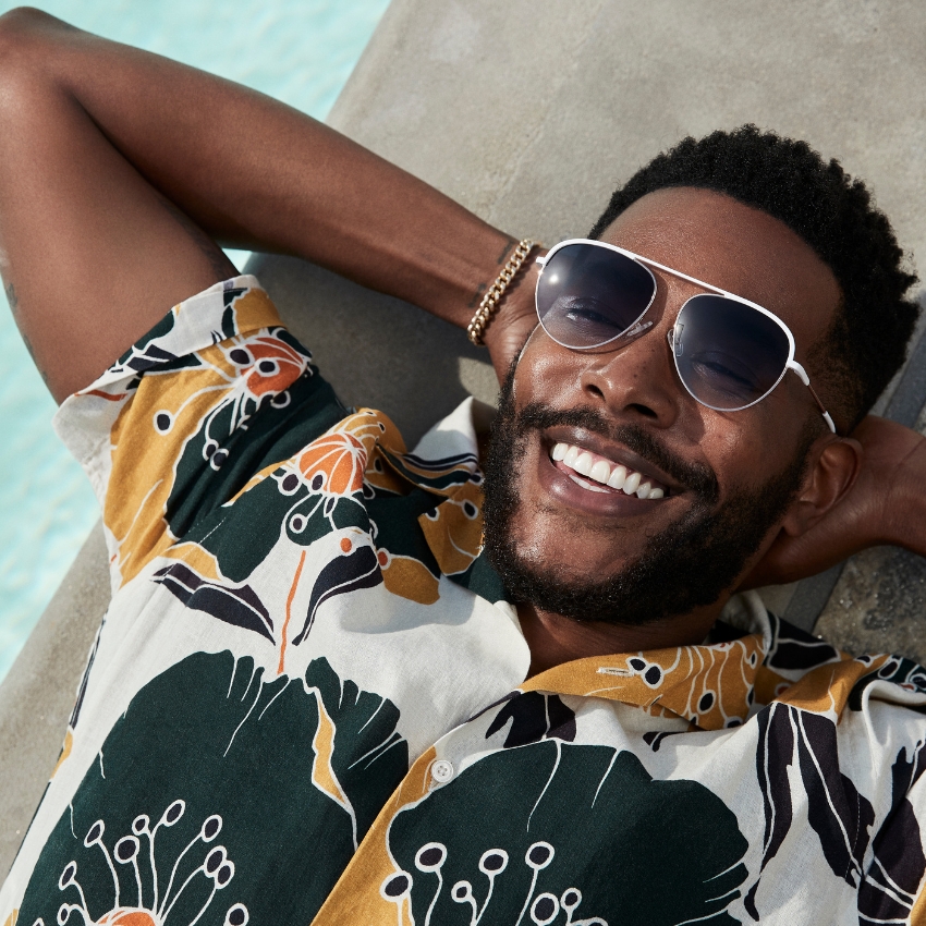 Man lounging by a pool, wearing aviator sunglasses and a floral-patterned, short-sleeve button-up shirt.