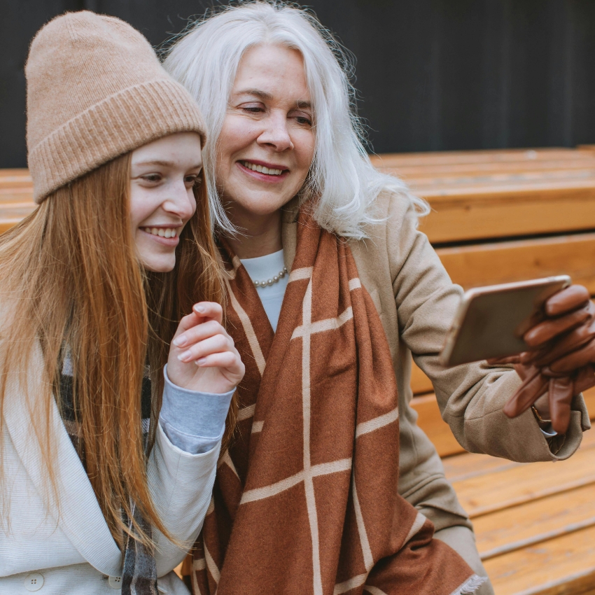 Two women in winter attire smile while looking at a smartphone.