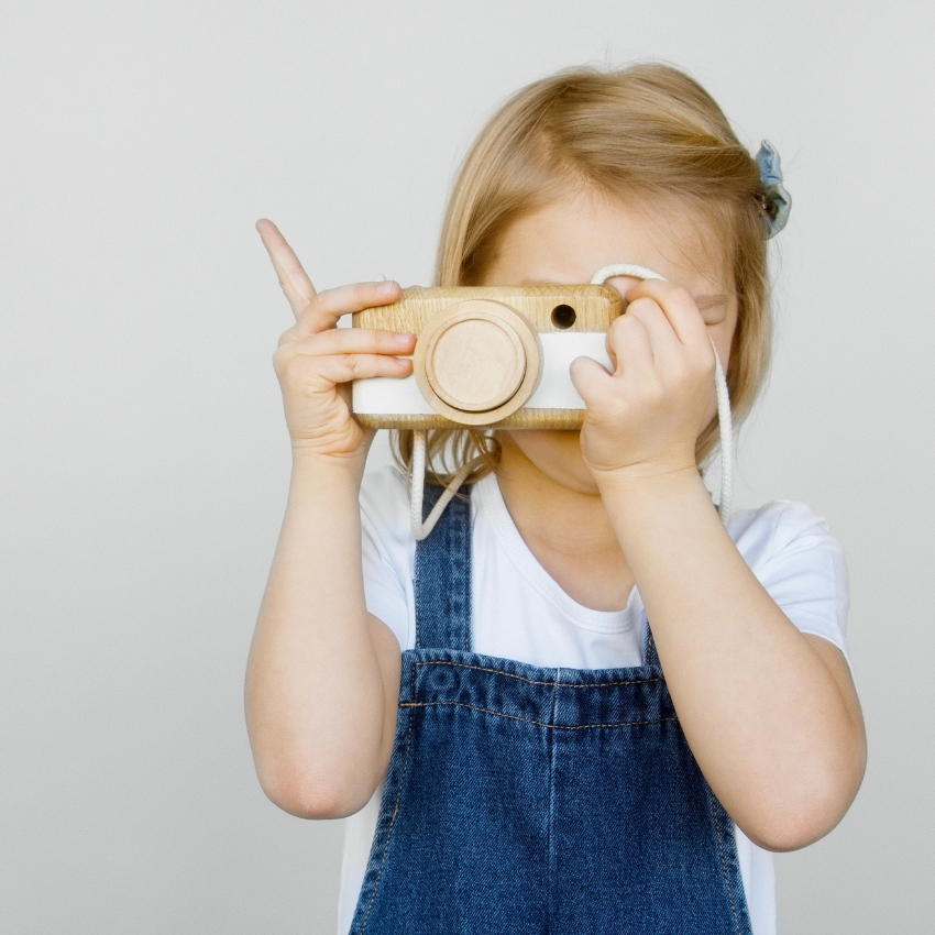 Child holding a wooden toy camera with a white strap, aiming as if taking a photo.