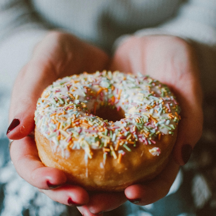 Donut with white icing and colorful sprinkles, held in both hands with red-painted nails.