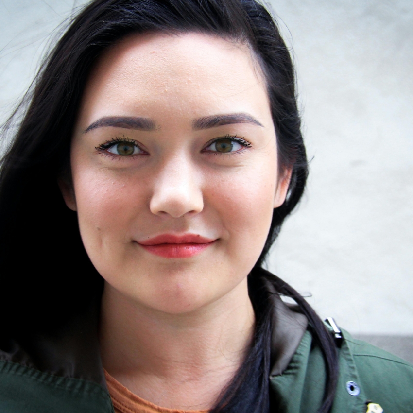 Woman with long black hair and green eyes, wearing a green jacket and a light brown top, smiling.