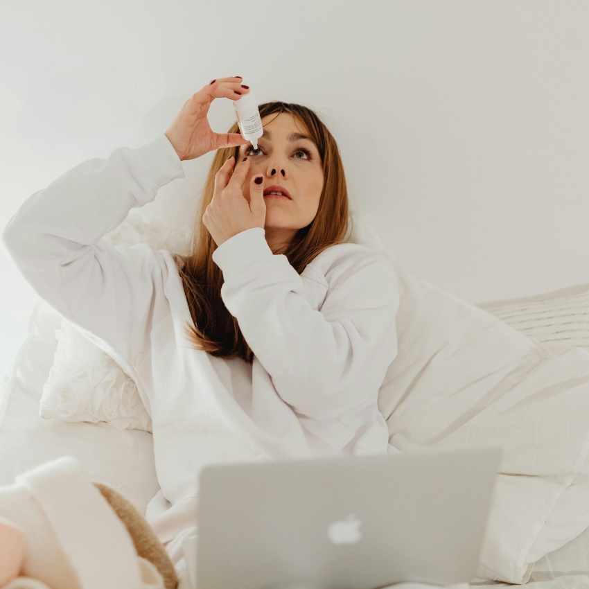 Woman applying eye drops while sitting in bed with a laptop.