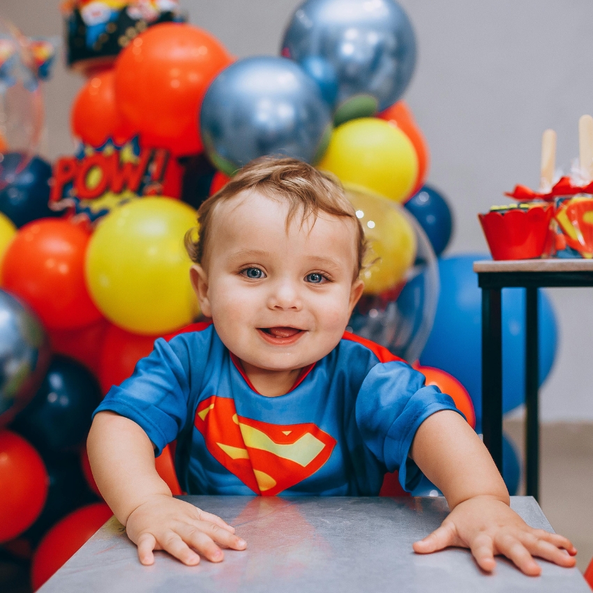 Baby wearing a blue Superman shirt with a red and yellow "S" symbol, in front of colorful balloons.