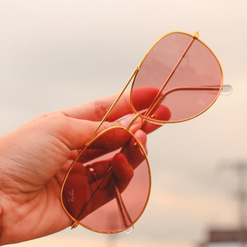 Gold-framed aviator sunglasses with pink lenses held by a hand. Small "Ray-Ban" logo on the lens.