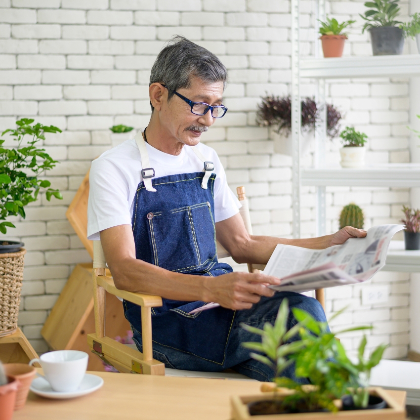 Senior man wearing denim apron reading a newspaper in a room with indoor plants.