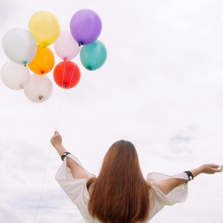 Person with long hair holding colorful balloons against a cloudy sky background.