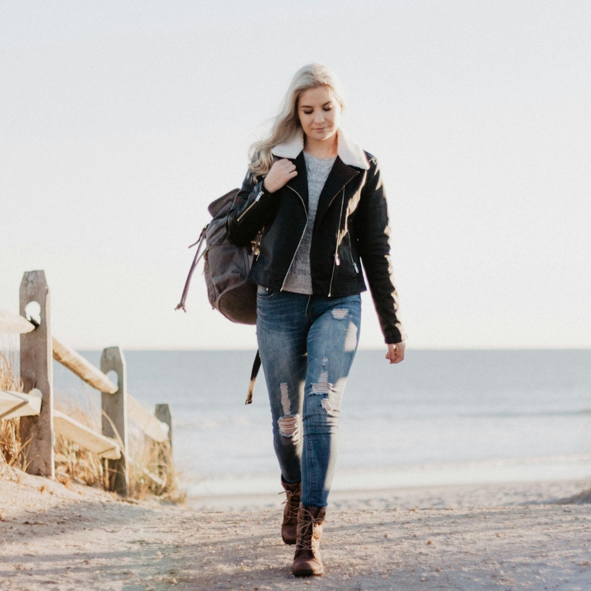 Woman wearing a black leather jacket, grey top, distressed jeans, and brown boots, carrying a large backpack.