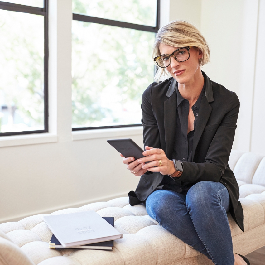 Woman holding an e-reader, seated on a couch with notebooks and a pen beside her.