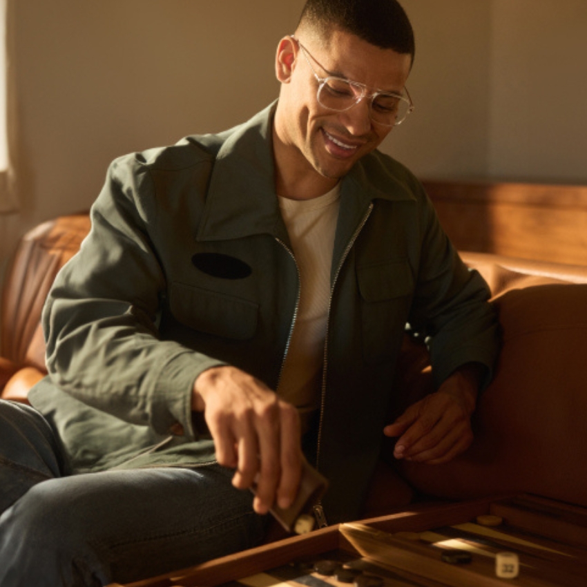 Person smiling while playing a game of backgammon, wearing a green jacket and eyeglasses.