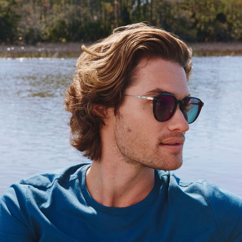 Man wearing round sunglasses with dark lenses and a blue t-shirt against a serene water background.