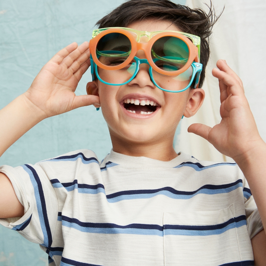 Child wearing two sets of oversized glasses, one orange and another blue, over a striped shirt.